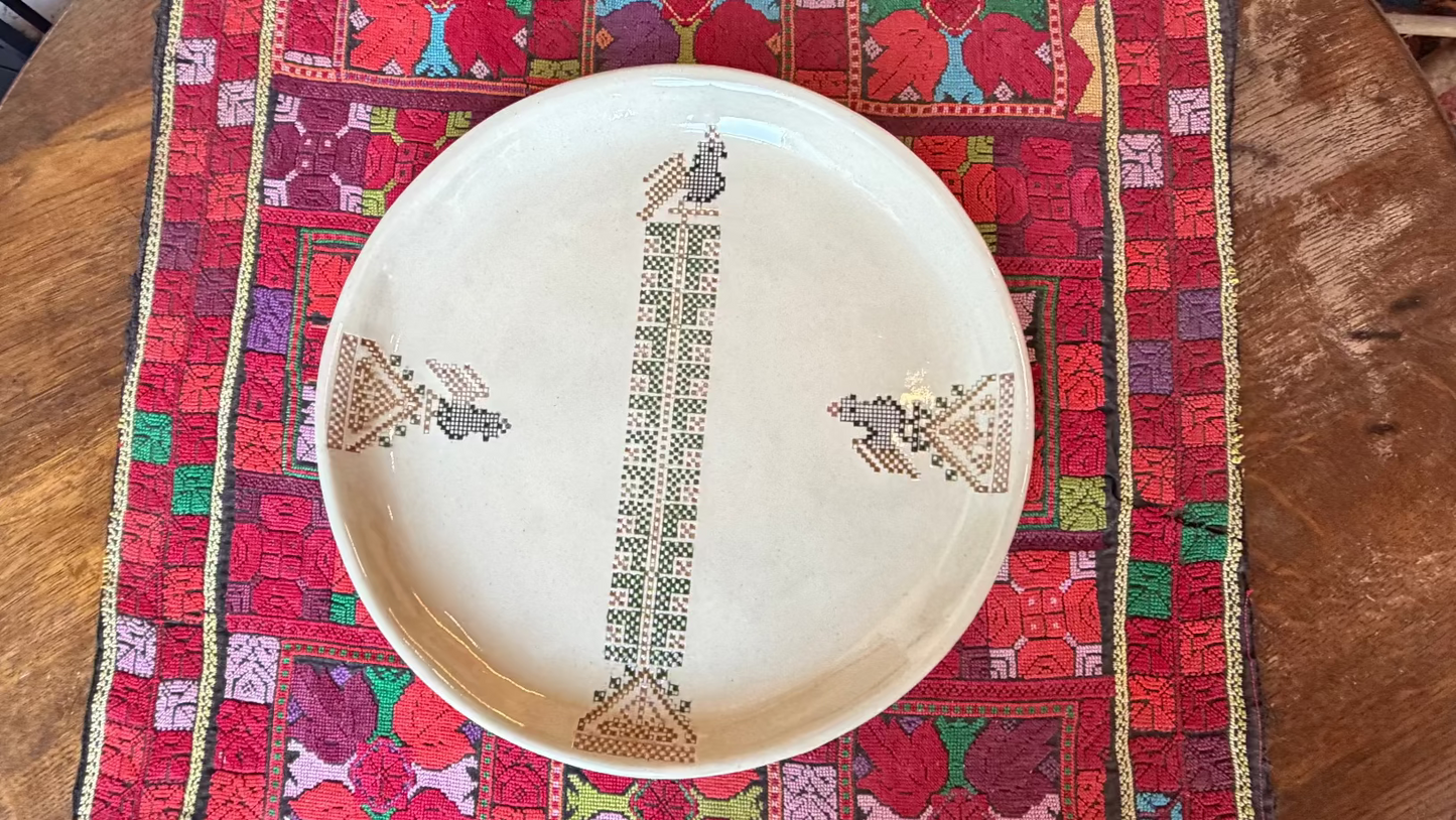 A round beige ceramic plate with traditional geometric and floral patterns, displayed on a colorful Palestinian embroidered textile over a wooden table.