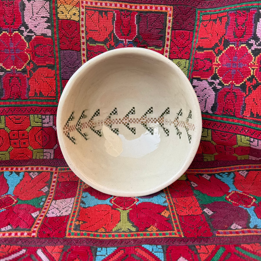 A small off-white ceramic bowl with a black and green geometric Palestinian embroidery pattern of a palm tree on the inside, displayed on a vibrant Palestinian textile background.