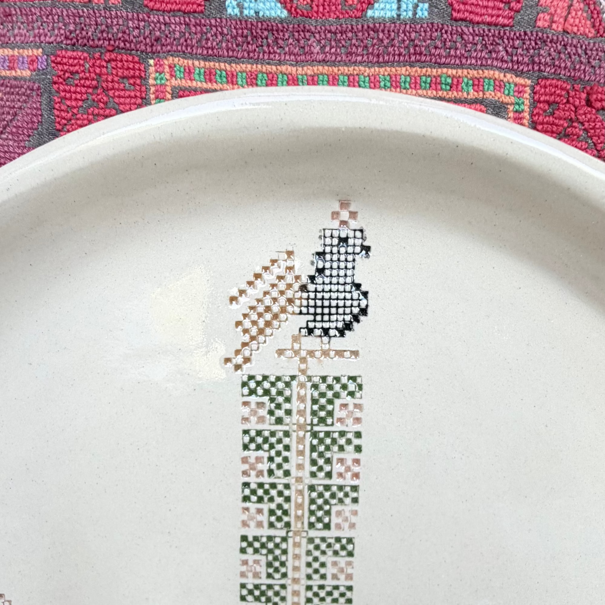 A close-up of a beige handmade ceramic plate featuring a hand-painted geometric pattern  of a rooster in brown, green, pink, and black, set against a colorful patterned Palestinian textile background.