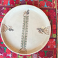 A round beige ceramic plate with traditional geometric and floral patterns, displayed on a colorful Palestinian embroidered textile over a wooden table.