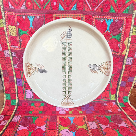 A round white ceramic plate with traditional geometric and floral patterns, displayed on a  colorful Palestinian embroidered textile. 