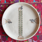 A round off-white ceramic plate with hand-painted geometric and floral patterns, displayed on a vibrant, multicolored Palestinian embroidered textile. 