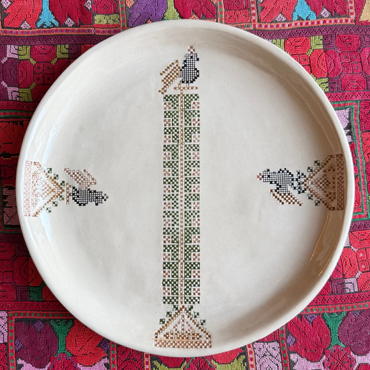 A round off-white ceramic plate with hand-painted geometric and floral patterns, displayed on a vibrant, multicolored Palestinian embroidered textile. 
